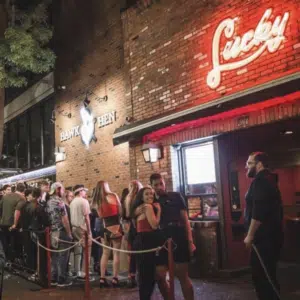 A group of people wait in line outside a brick building with neon "Lucky" sign at night.